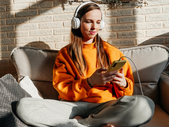 Eine Frau sitzt auf einem Sofa und hört über Handy und Kopfhörer einen Podcast.